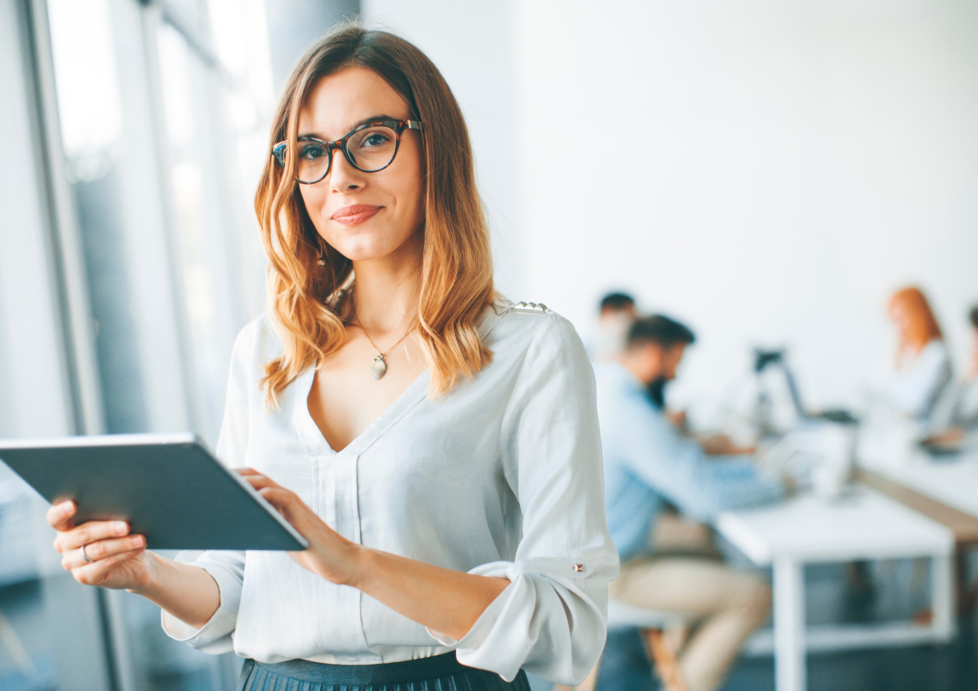 Smiling woman with glasses holding a tablet in a modern office with colleagues blurred in background.