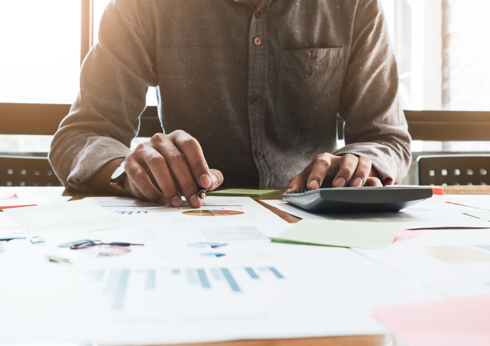 Person using a calculator and pen to analyze financial charts and documents on a desk.