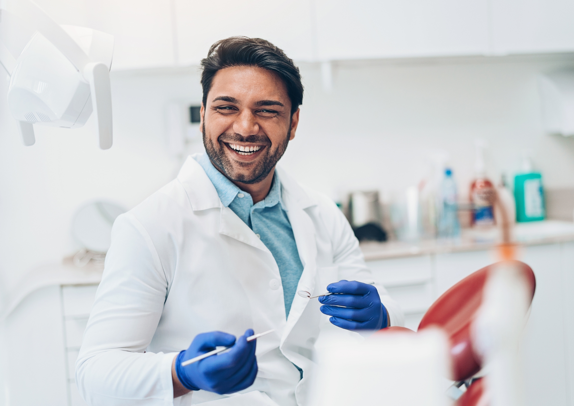 Smiling male dentist in white coat and blue gloves holding dental tools in a clinic.