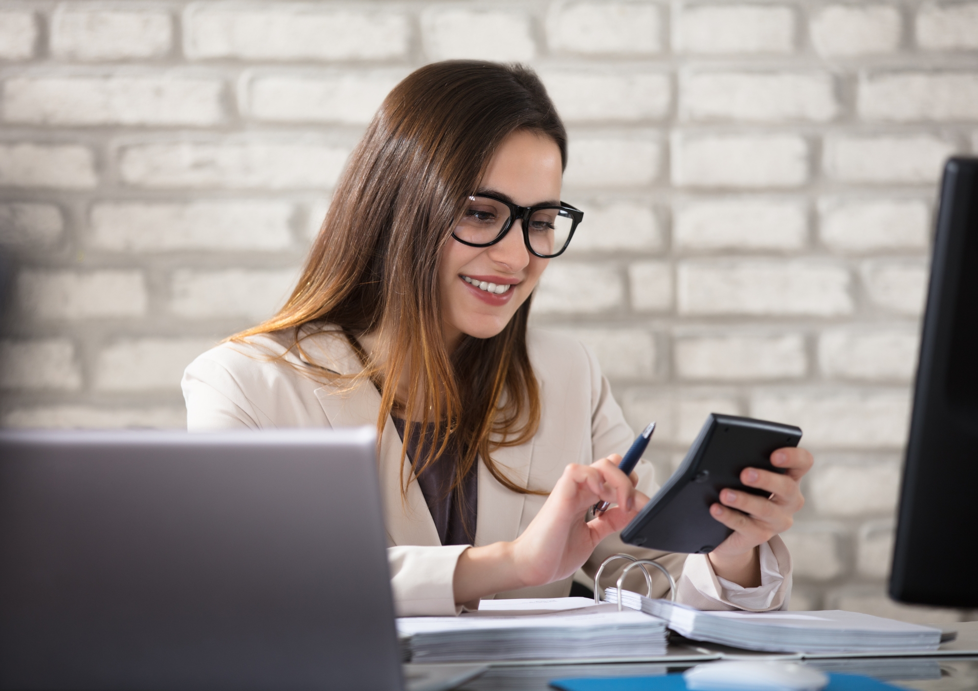 Smiling woman in glasses using a calculator at her desk with documents and a laptop