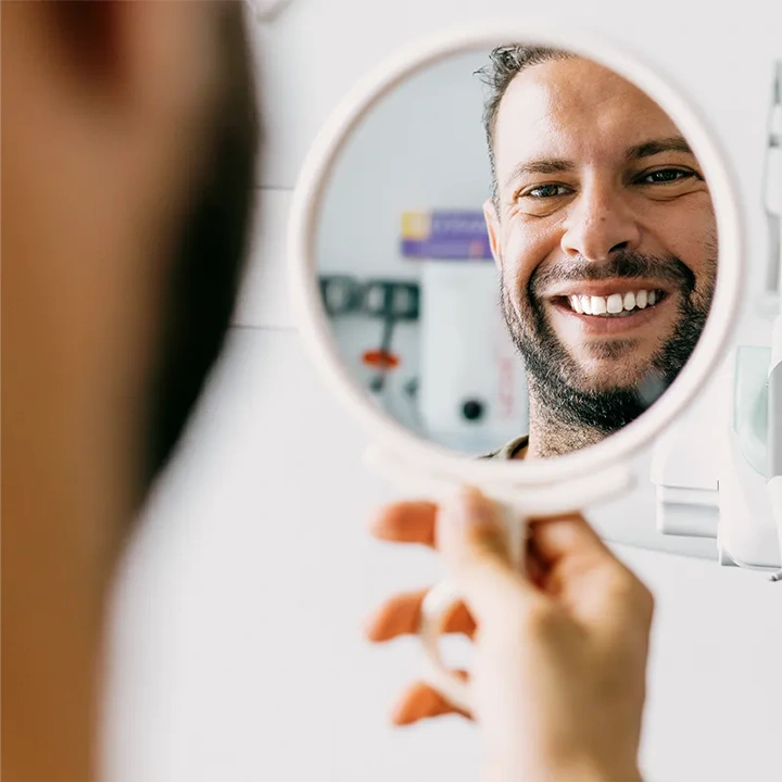 Man with beard smiling at his reflection in a round hand mirror indoors.
