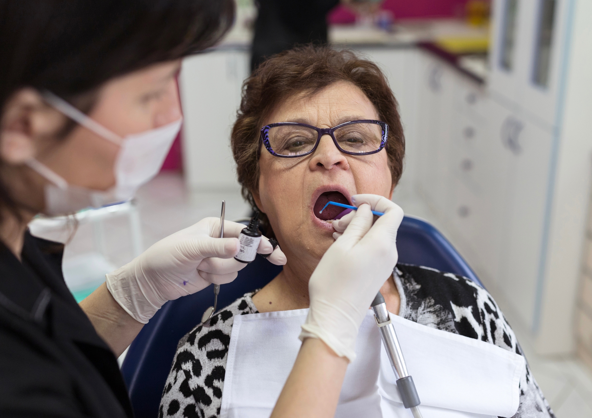 Dentist wearing gloves and mask examining older woman’s open mouth with dental tools.