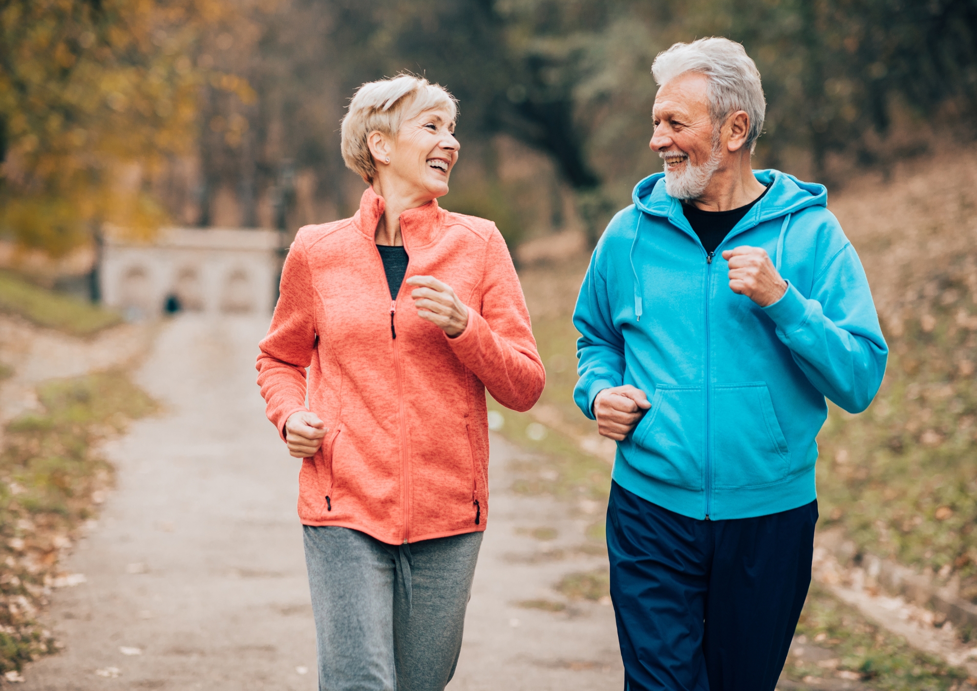 Smiling elderly couple jogging outdoors on a path in autumn wearing casual sportswear.