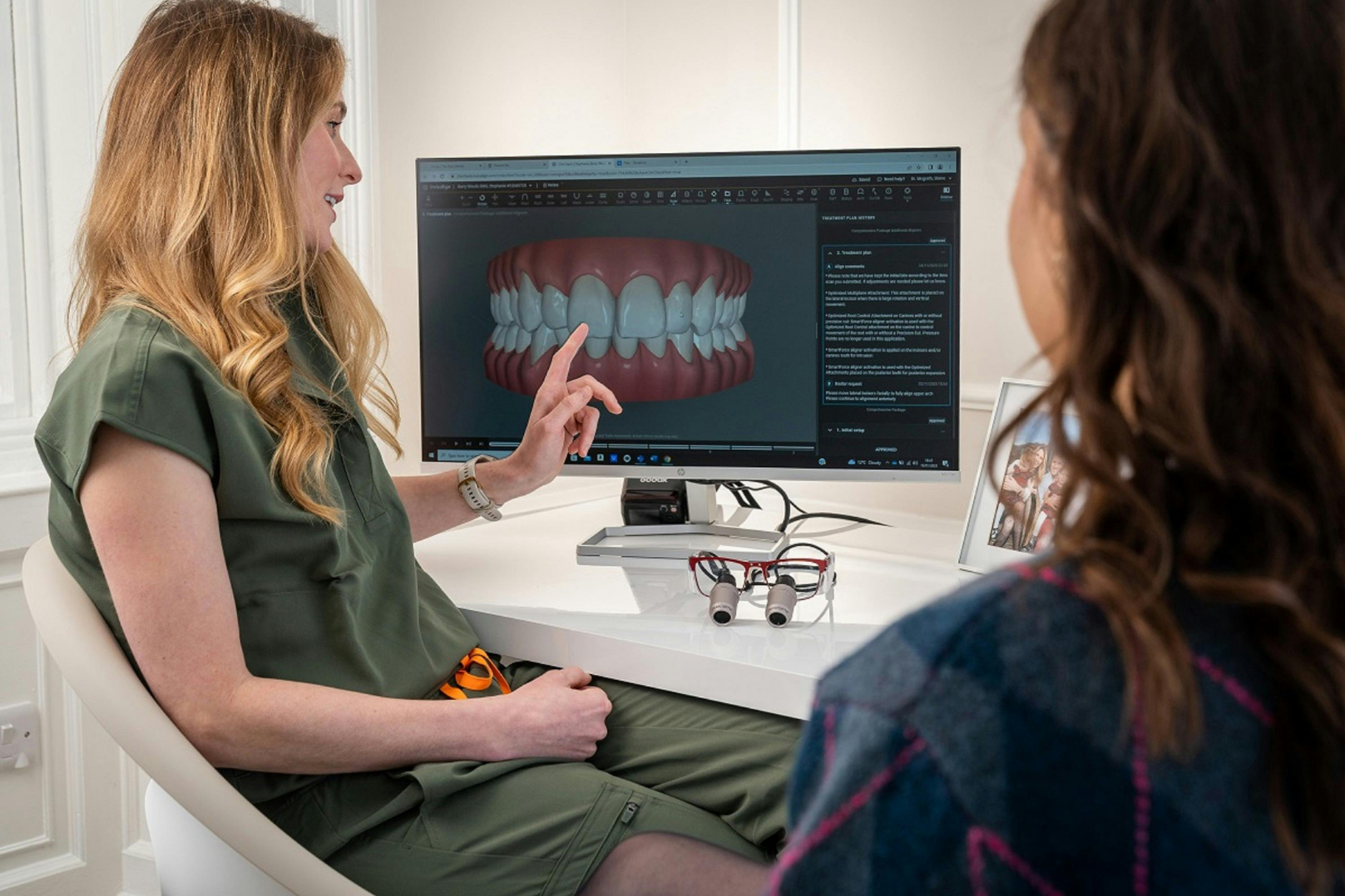Dental professional explains 3D teeth model on computer screen to patient in consultation room.