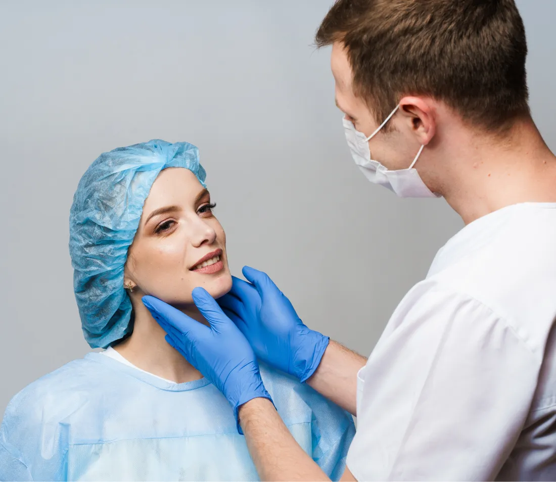 Doctor in gloves and mask examining female patient's face wearing surgical cap and gown.