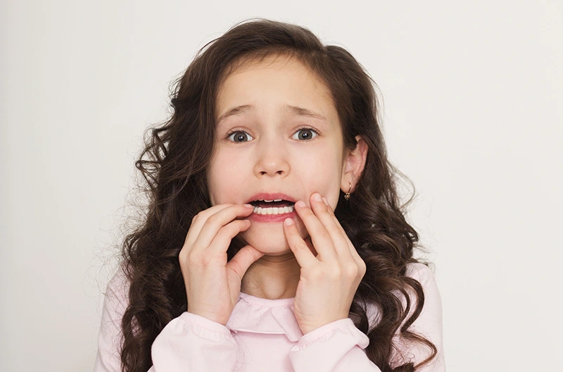 Worried young girl with curly hair biting nails against a plain background.