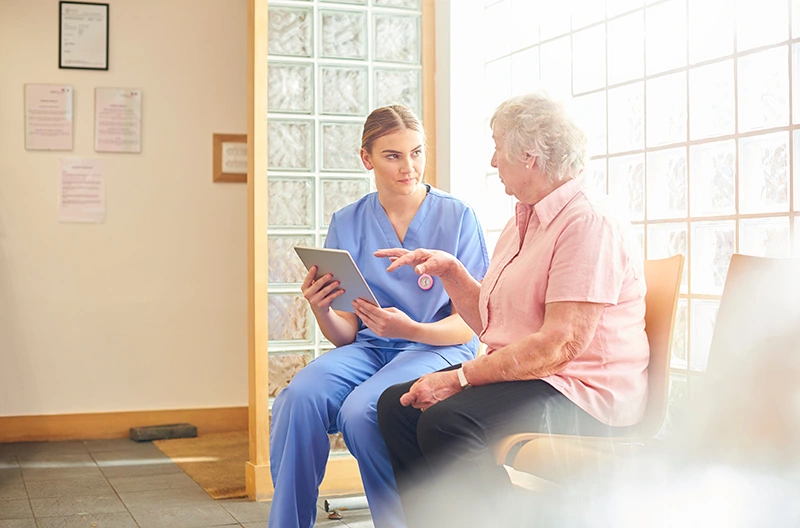 Nurse in blue scrubs speaking with elderly woman in pink shirt in a bright waiting room.