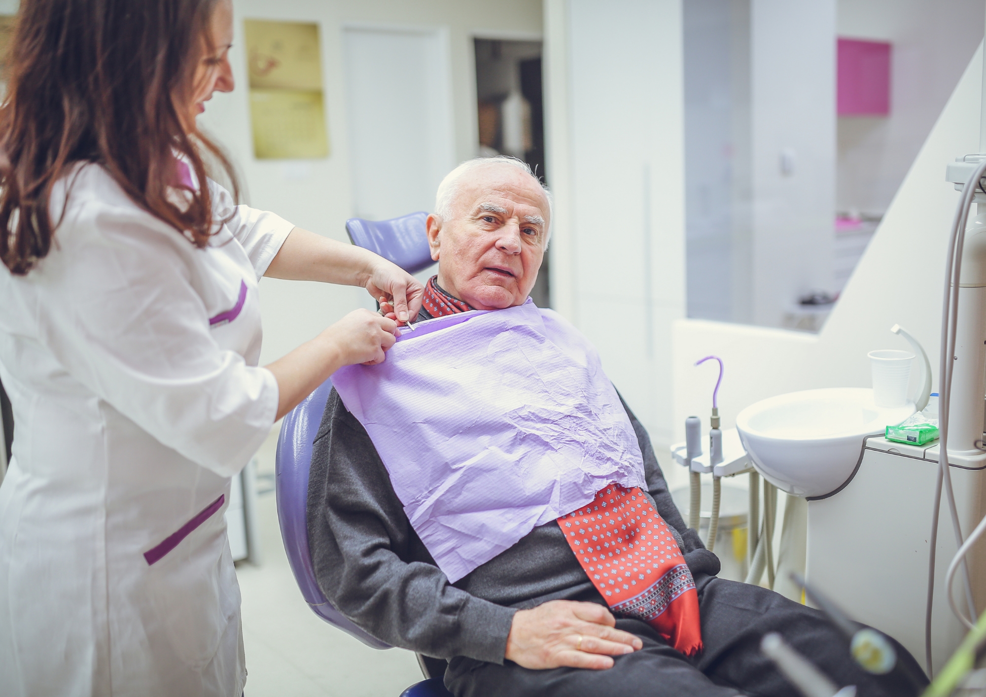 Dentist placing a dental bib on an elderly man seated in a dental chair.