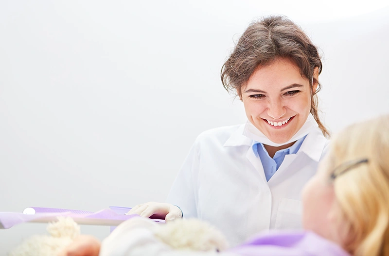 Smiling female dentist in white coat looking at a patient lying in a dental chair.