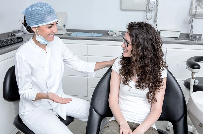 Smiling female dentist talking to a seated female patient in a modern dental office.