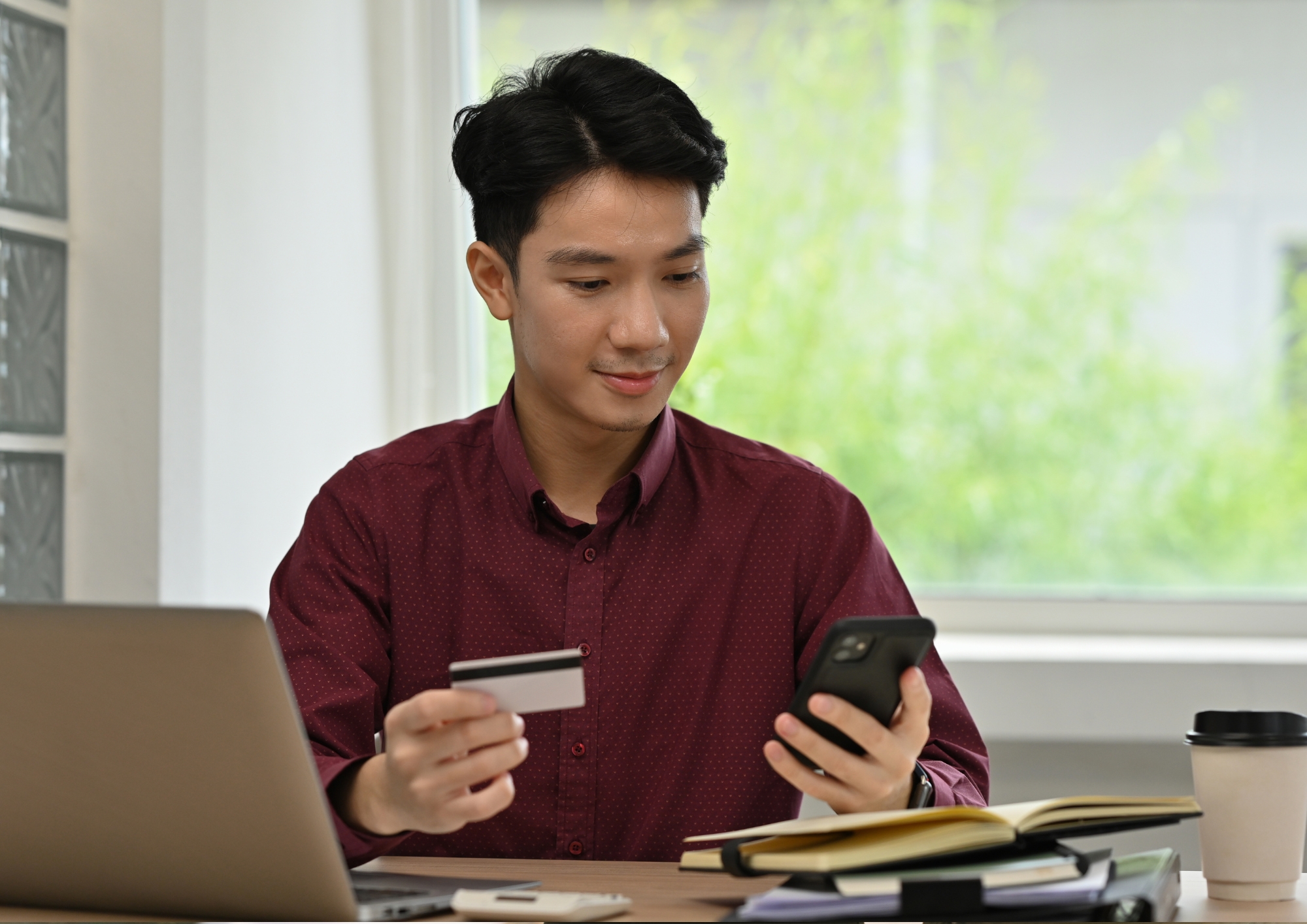 Man holding credit card and smartphone, sitting at desk with laptop, books, and coffee cup.