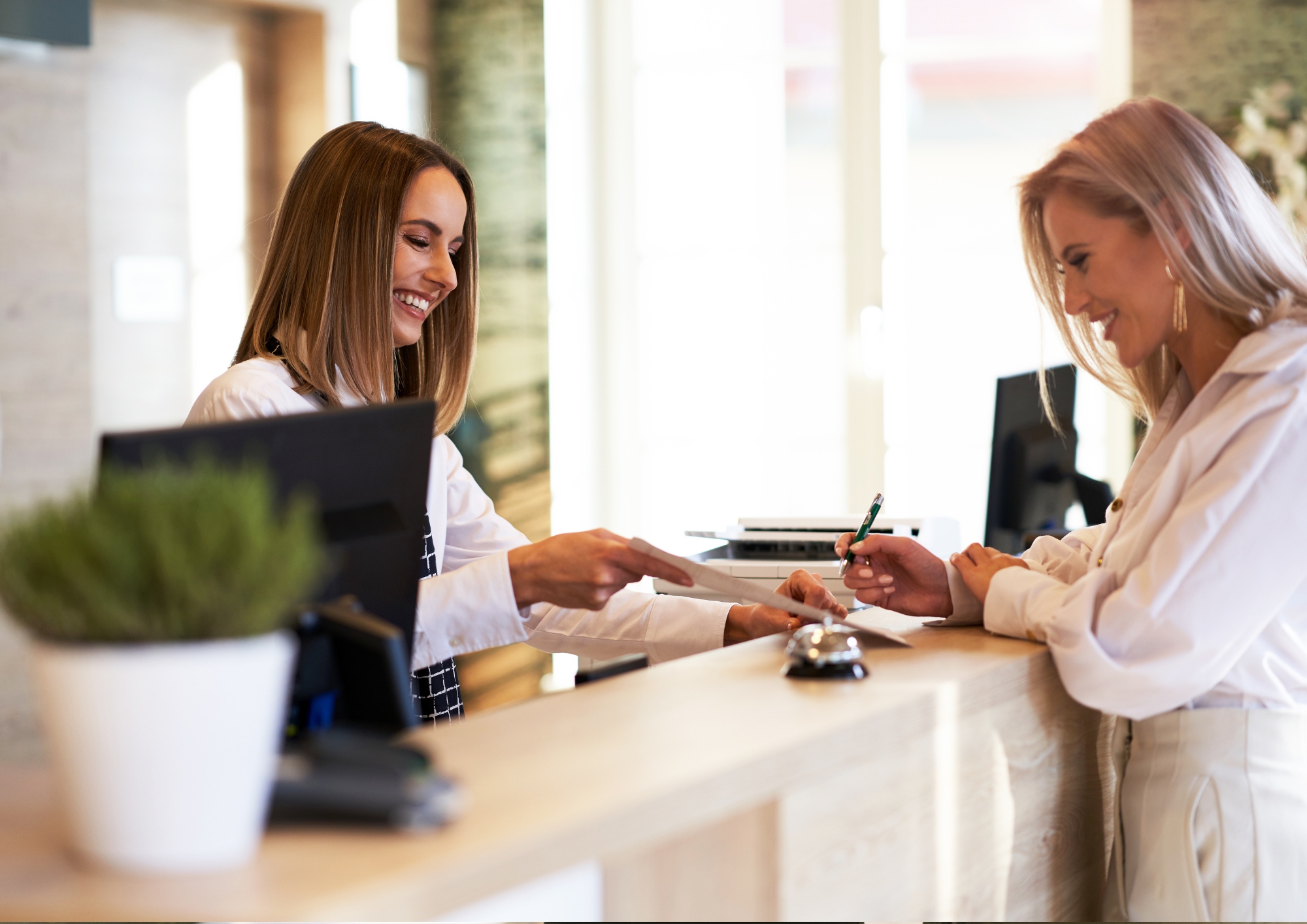 Smiling receptionist handing paper to a guest signing documents at a hotel front desk.