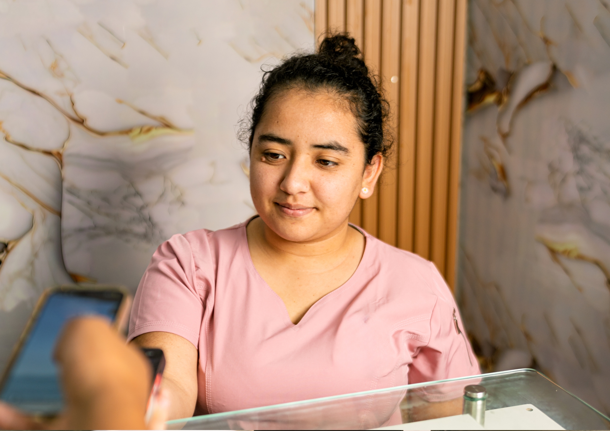 Woman in a pink shirt looks at a smartphone held by another person across a glass counter.