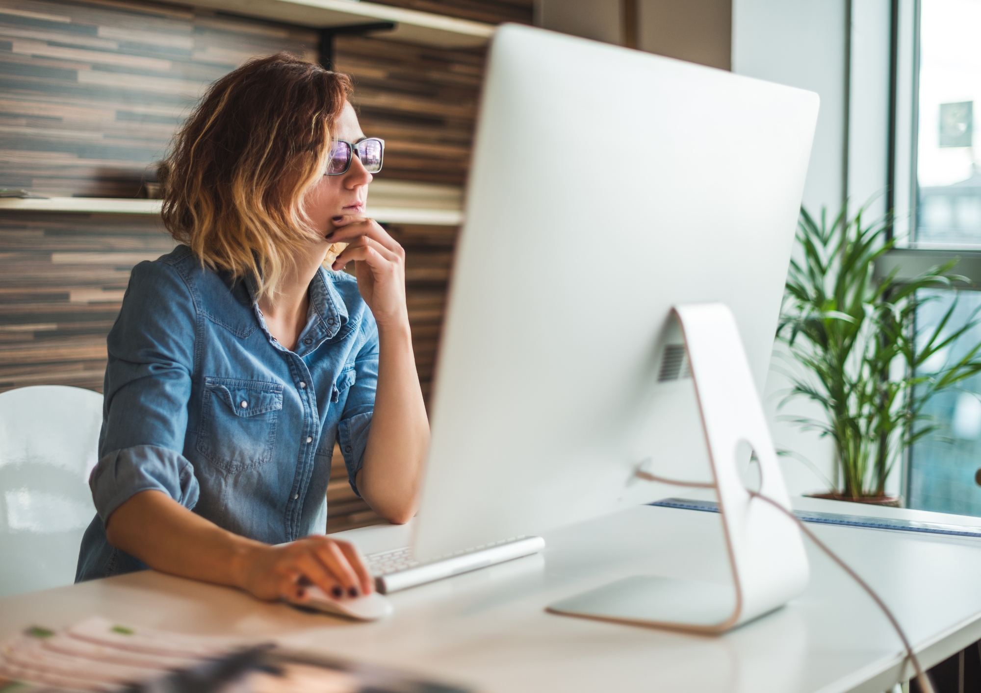 Woman in glasses wearing denim shirt working on desktop computer in bright modern office.