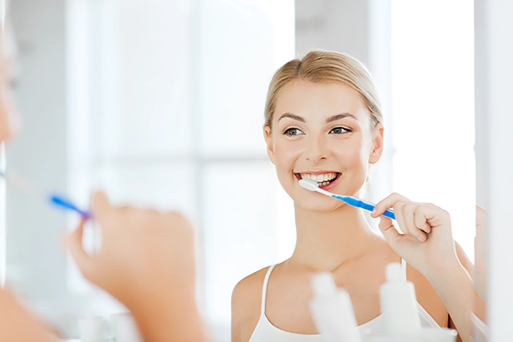 Smiling woman brushing her teeth while looking at a mirror in a bright bathroom.