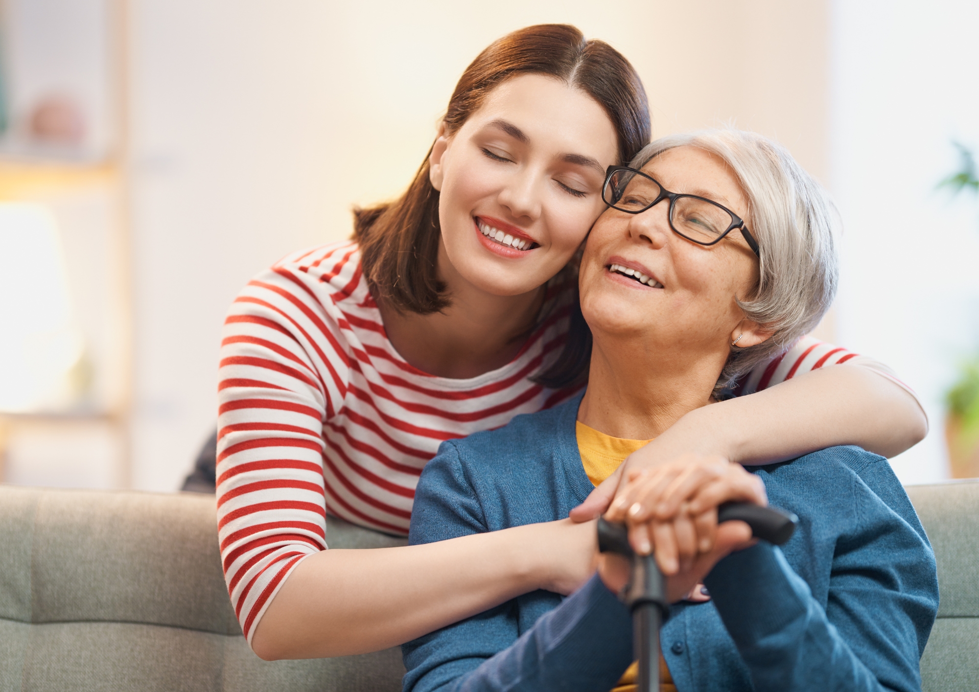 Young woman hugs smiling elderly woman holding a cane, sitting on a couch in a warmly lit room.