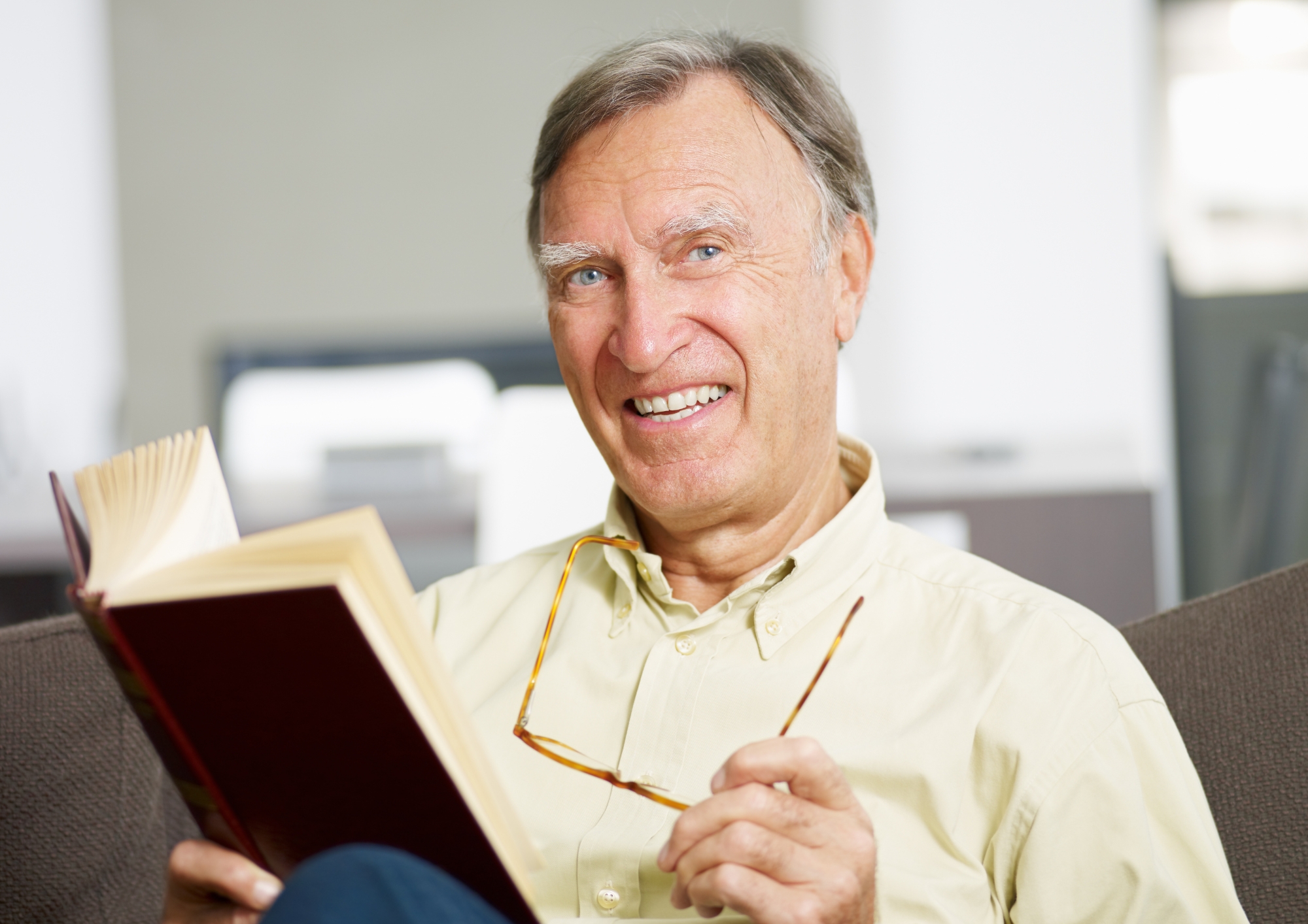 Smiling elderly man holding glasses and reading a book indoors on a couch.