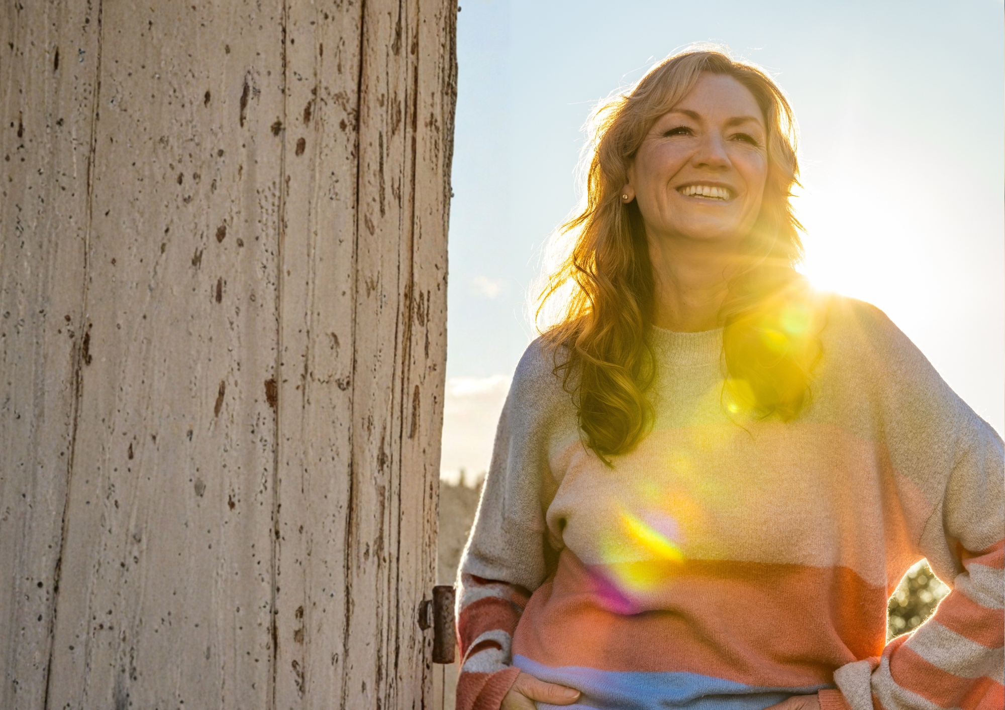 Smiling woman in a colorful sweater with sunlight flares behind her, standing by a textured wall.