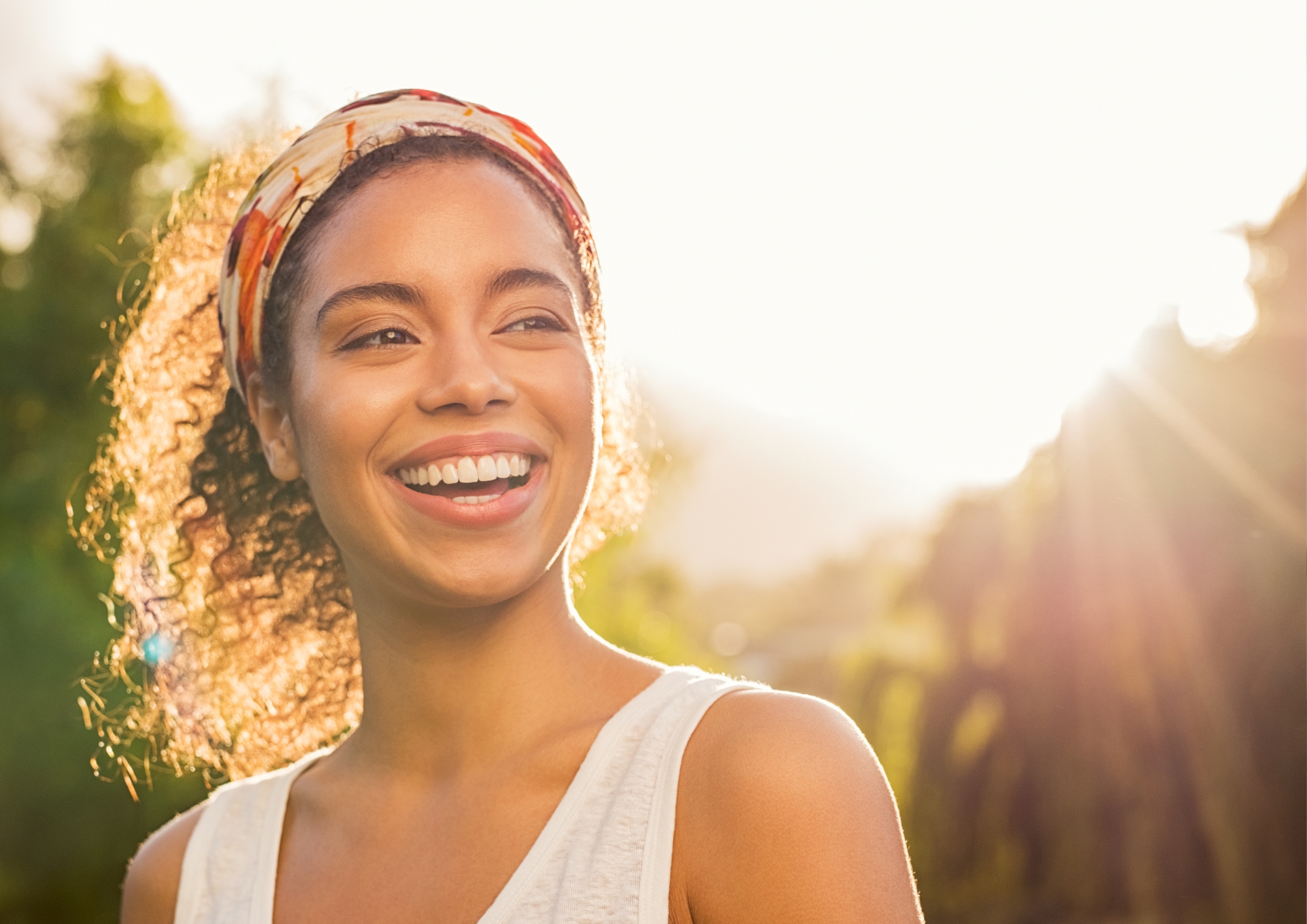 Smiling woman with curly hair and colorful headband outdoors with sunlight in background.