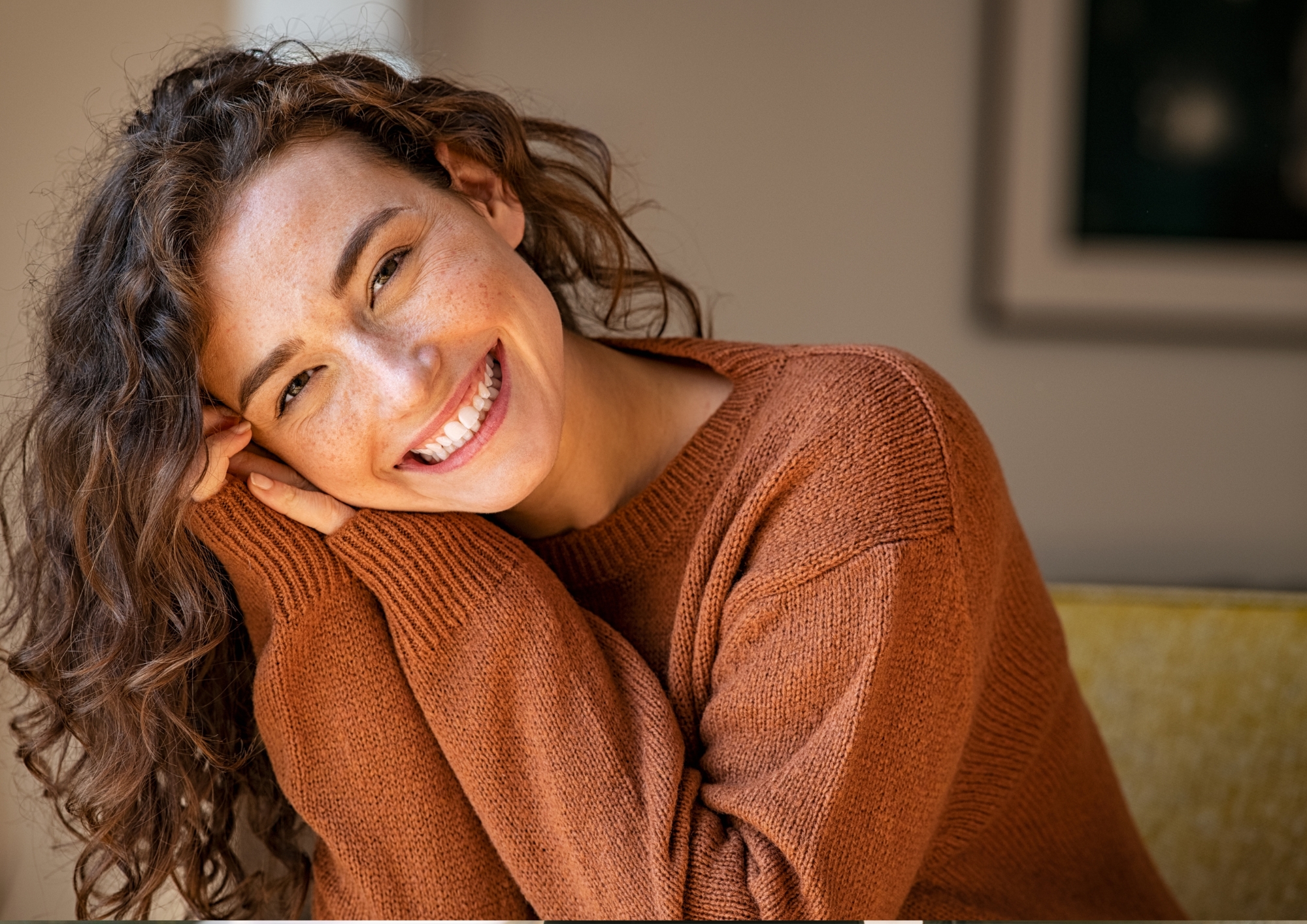 Woman with curly hair smiling, wearing an orange-brown sweater, resting her head on her hands.