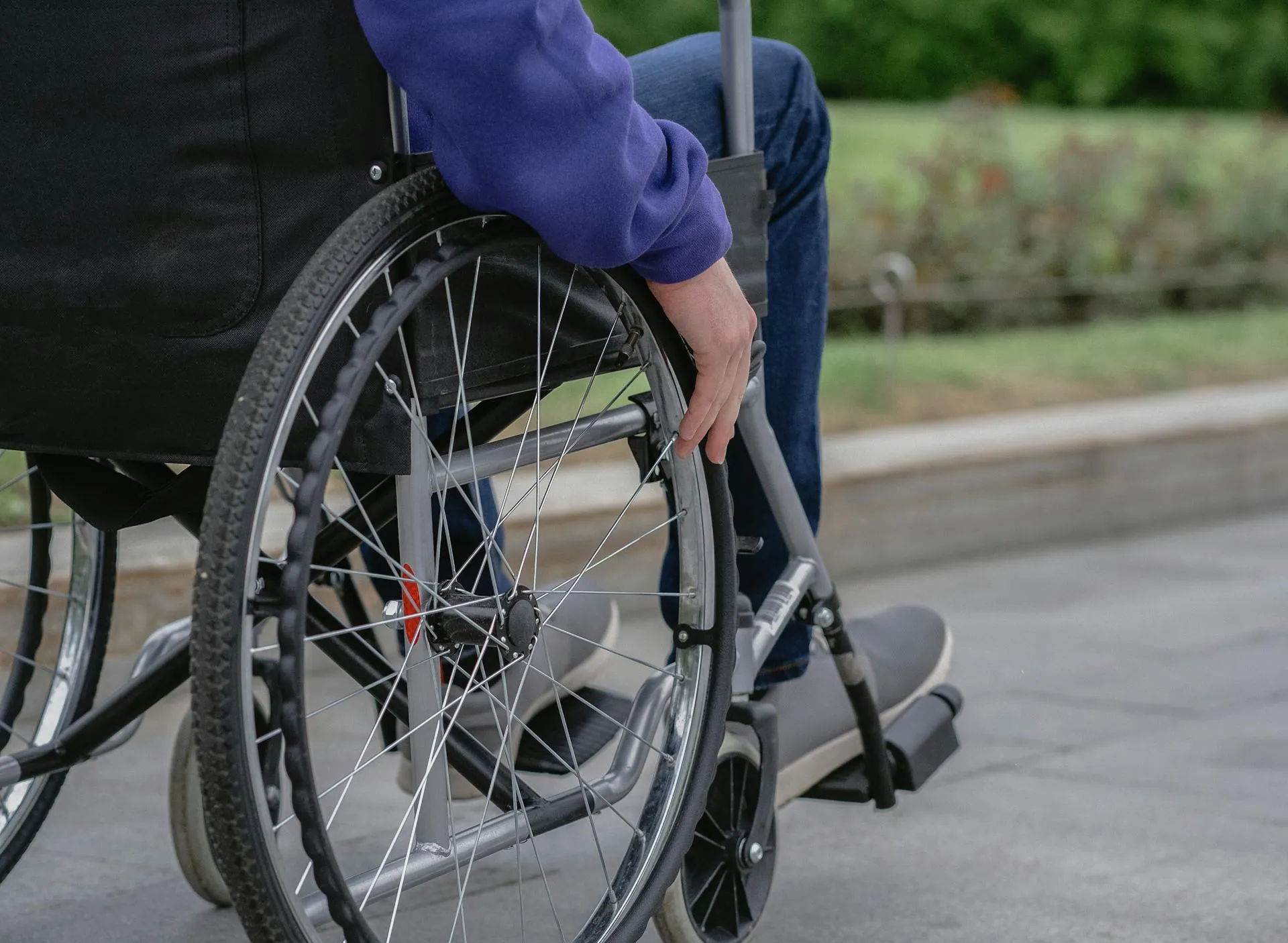 Person in wheelchair wearing blue jeans and gray shoes outdoors on a paved path.