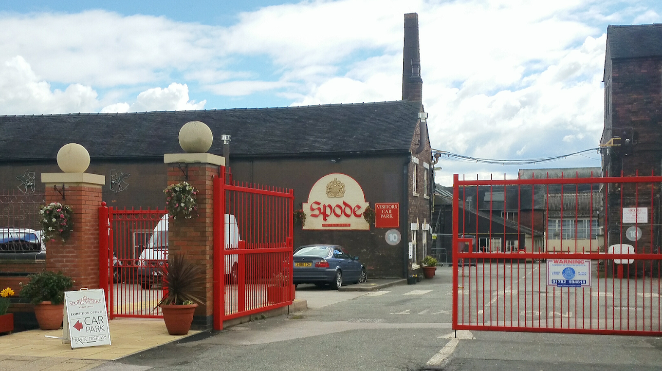 Entrance of Spode factory with red gates, parked cars, and signs for visitor car park and exhibition.