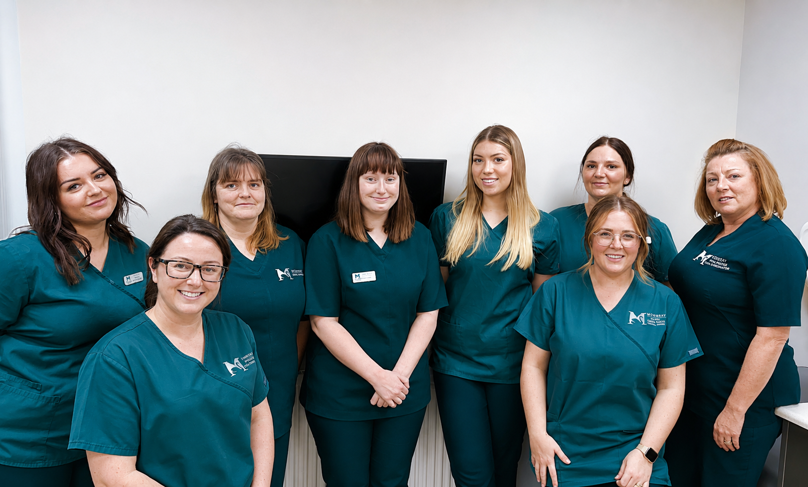 Group of eight women in teal medical scrubs standing and sitting indoors against a plain wall.
