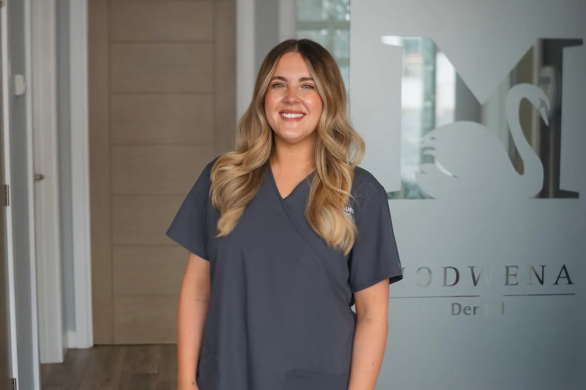 Smiling woman with long blonde hair wearing gray scrubs standing in front of a frosted glass door.