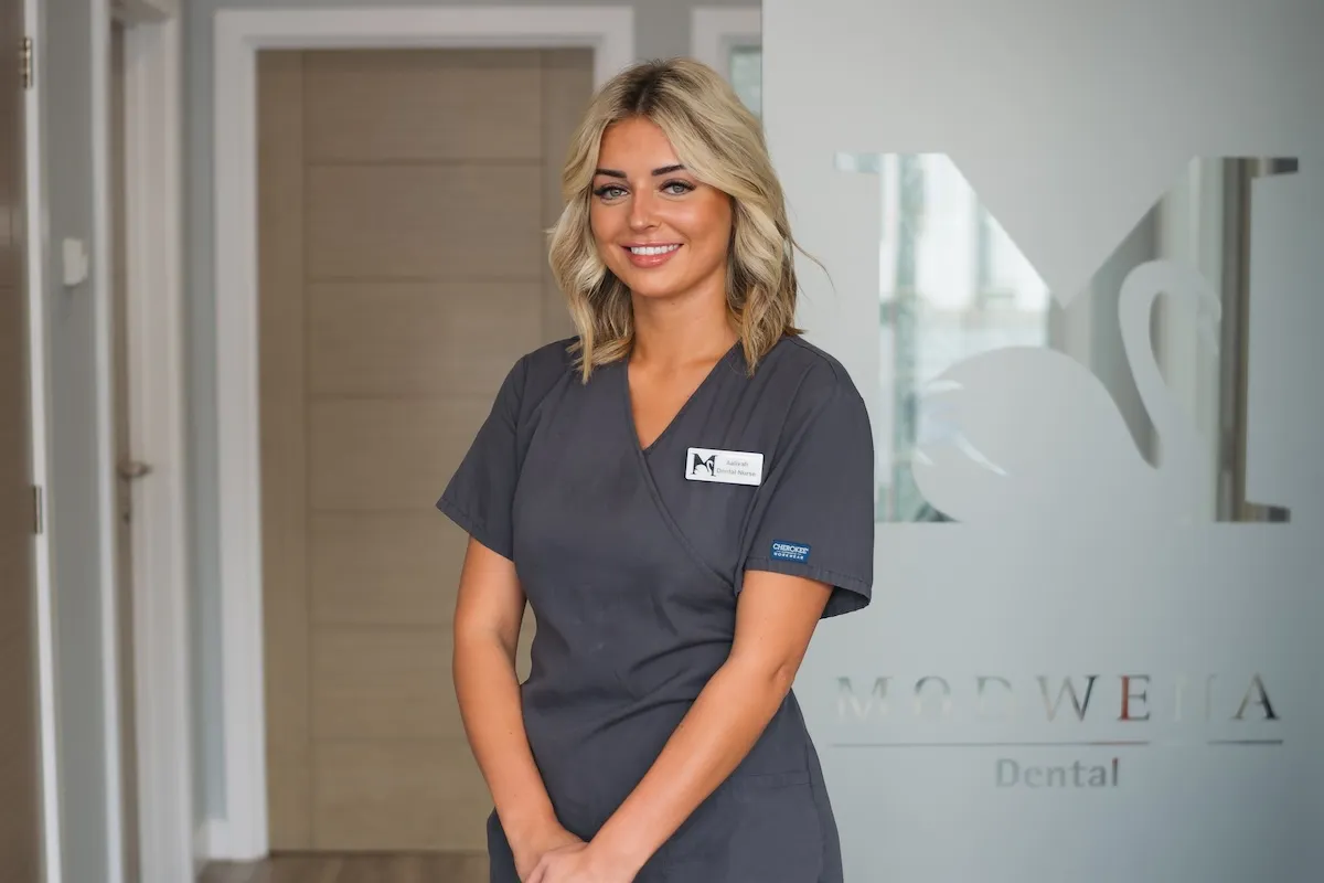 Smiling dental nurse in dark scrubs standing in a dental clinic reception area.