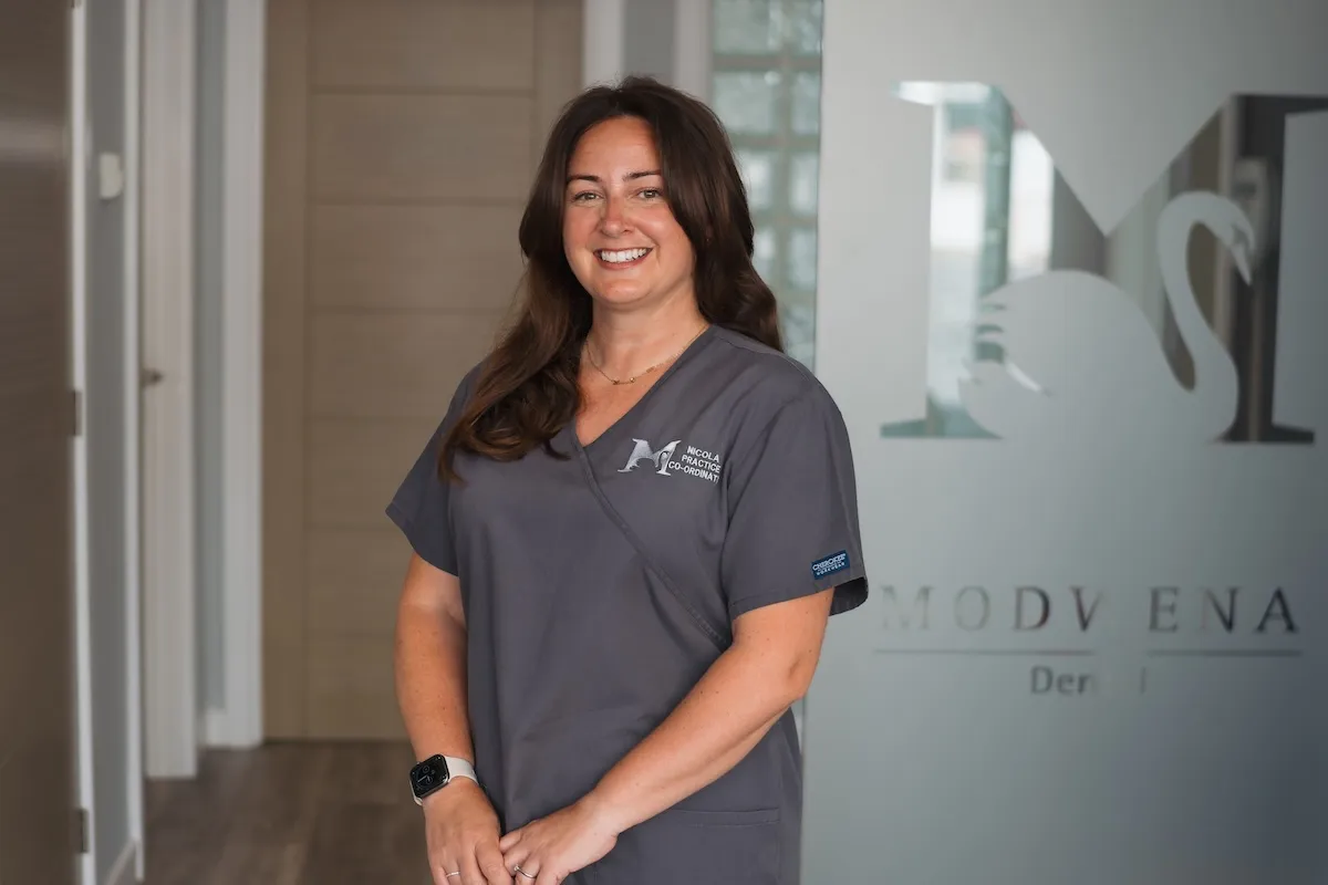 Smiling woman in gray scrubs standing in a dental office hallway with a frosted glass door.