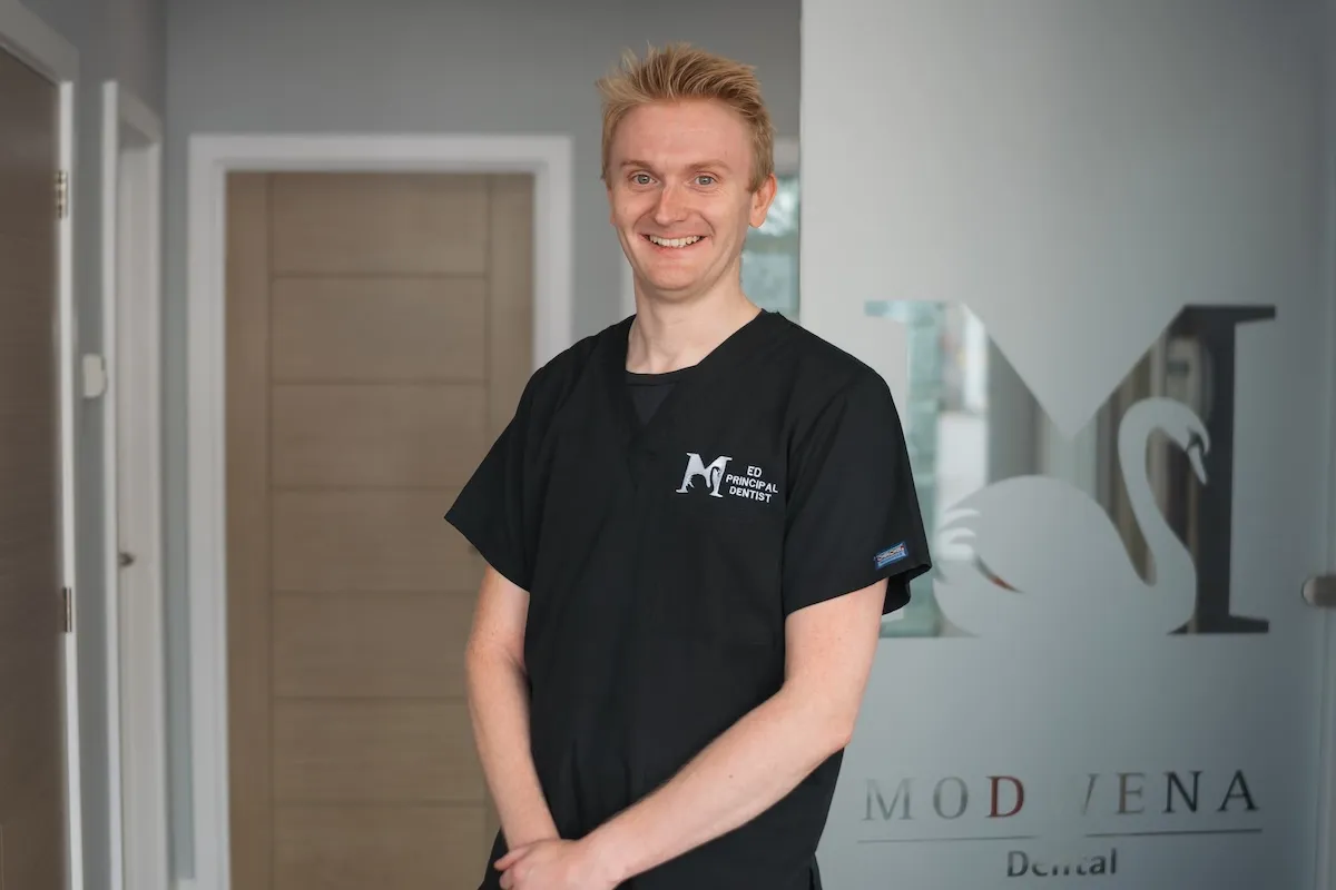 Smiling man in black scrubs with Modwena Dental logo standing inside a dental clinic.
