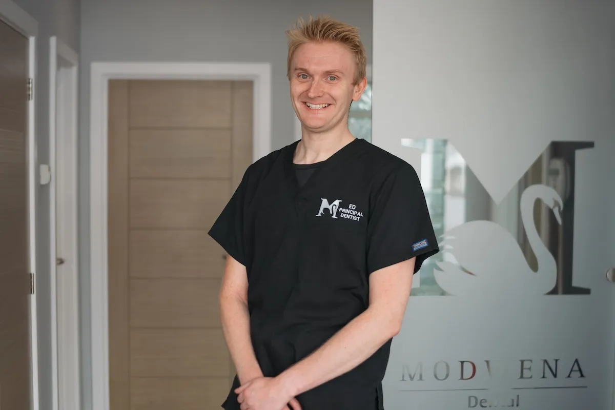 Smiling man wearing black scrubs stands in front of a door and a glass panel with a swan logo.