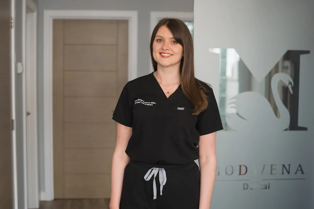 Smiling woman dental therapist in black scrubs standing indoors with a logo on glass behind her.