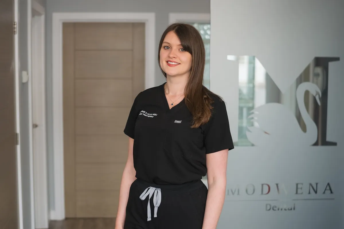 Smiling woman in black scrubs standing in a dental office near a frosted glass door with a swan logo.