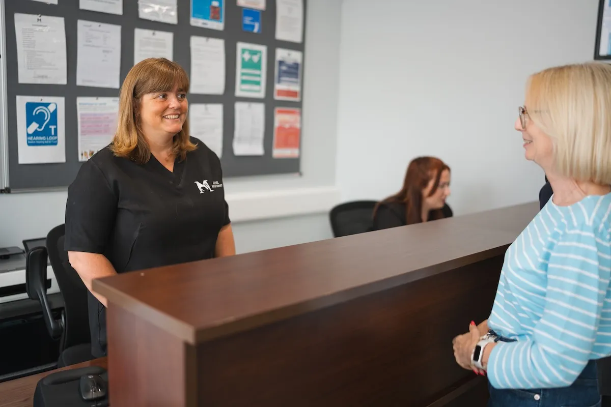 Dental receptionist smiling behind a desk talking with a woman wearing glasses and a striped shirt.