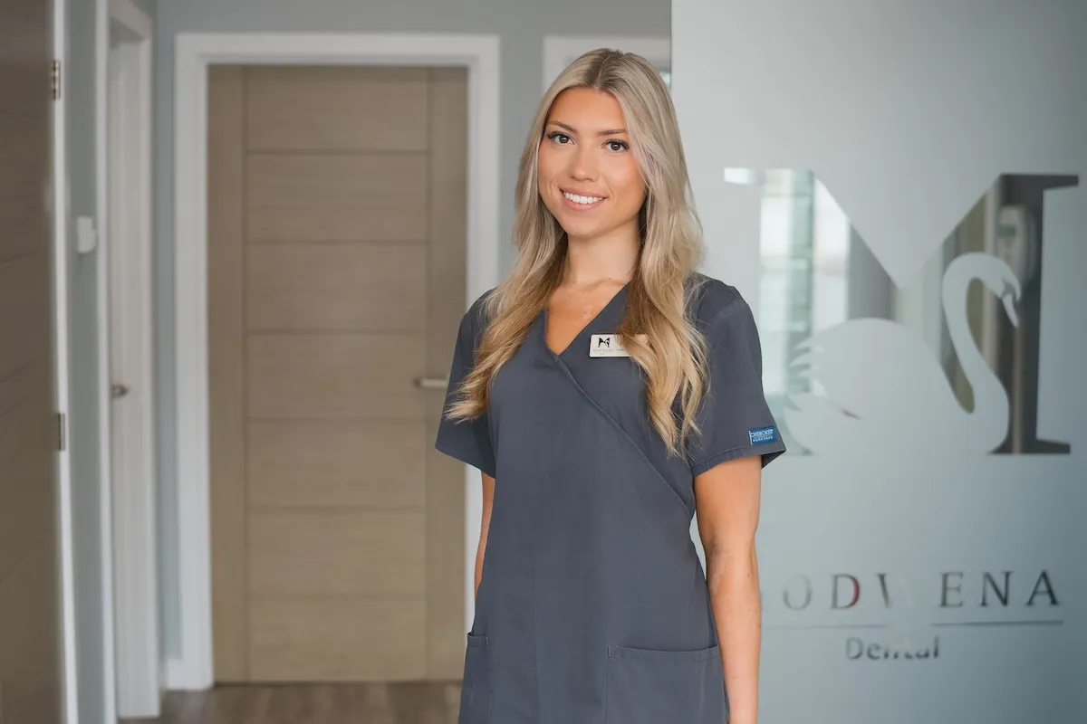 Smiling dental professional in gray scrubs standing inside a dental office near a glass door.