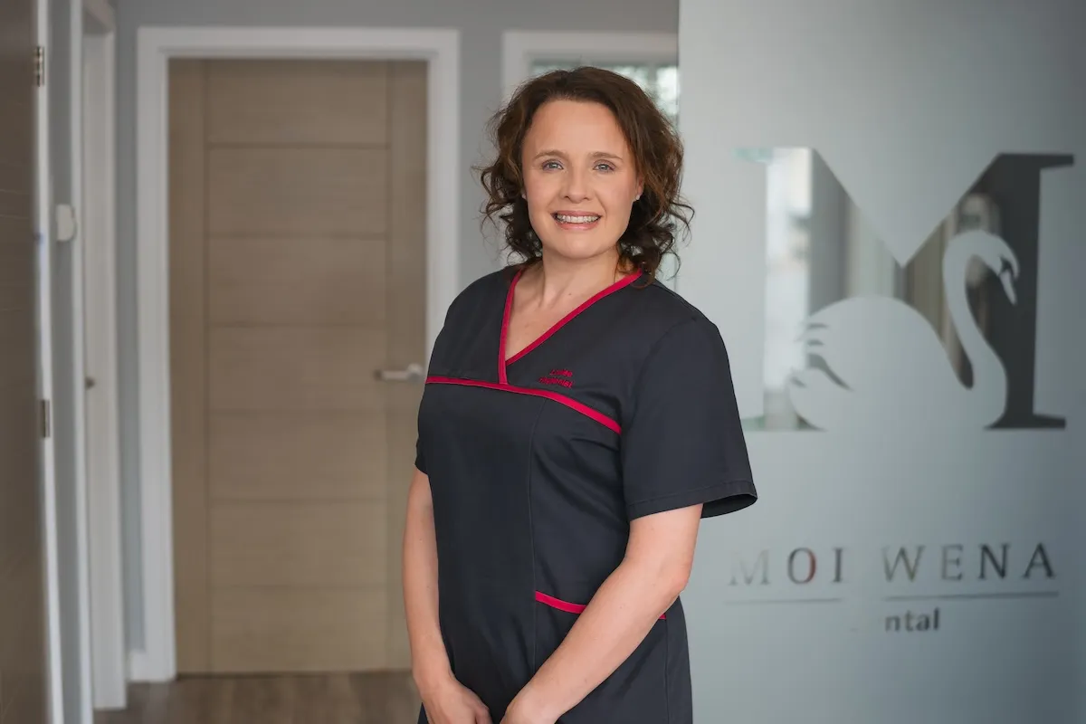 Smiling woman in black dental uniform standing in a modern clinic with a glass wall logo behind her.