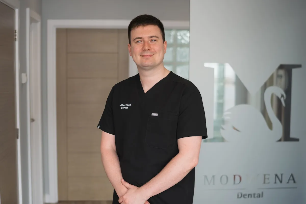 Smiling man in black dental scrubs standing inside a dental clinic with Modwena Dental sign behind.
