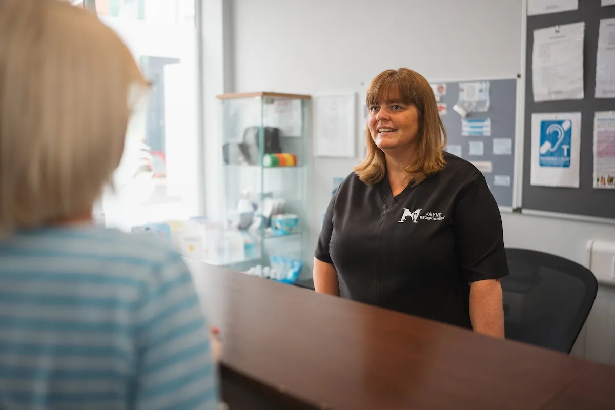 Receptionist named Jayne smiling behind a desk talking to a woman in a striped shirt.