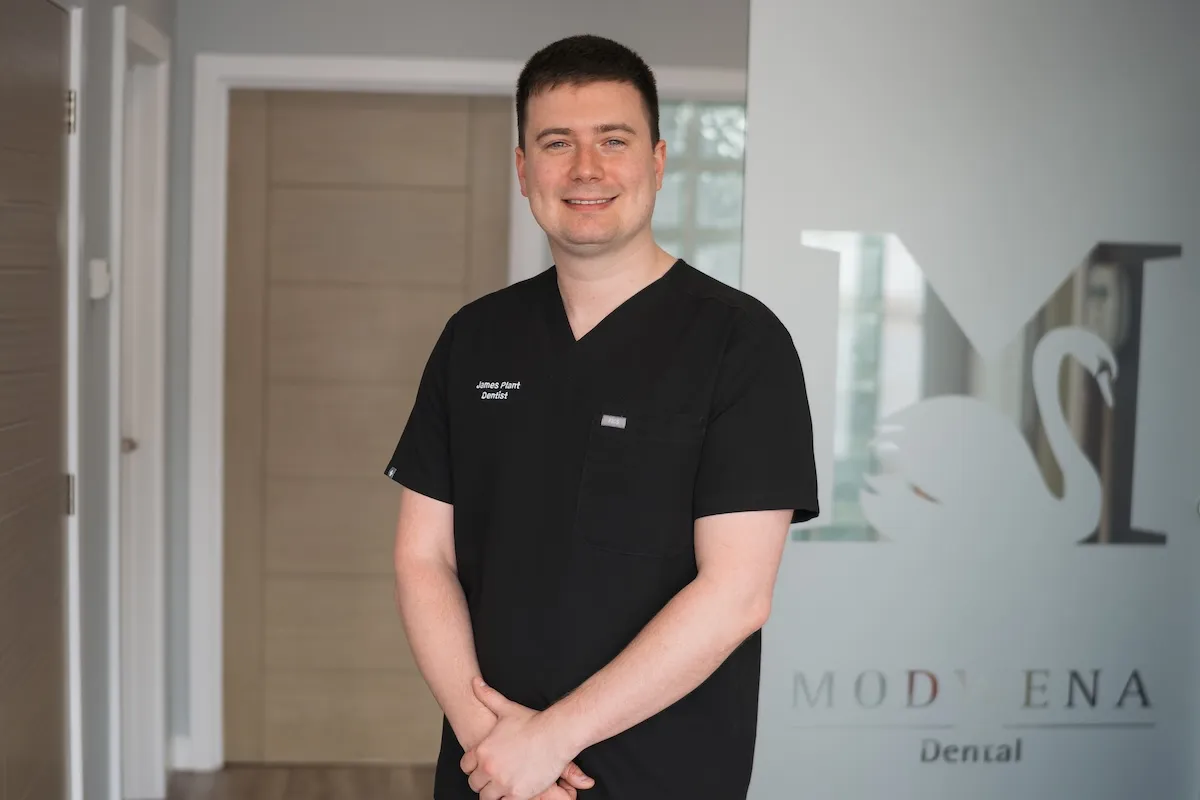Smiling man in black scrubs with James Plant Dentist badge stands inside a dental clinic.