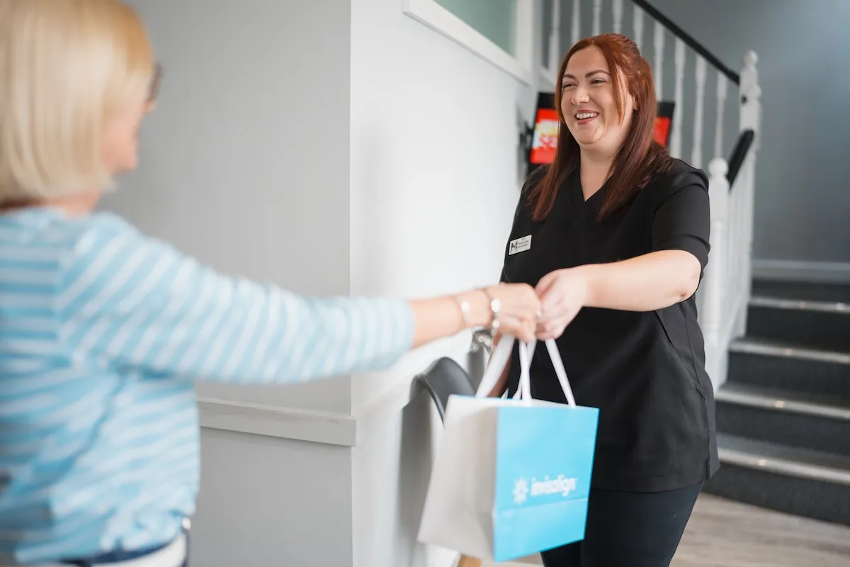 Smiling woman in black gives a white and blue Invisalign bag to a blond woman in striped shirt.