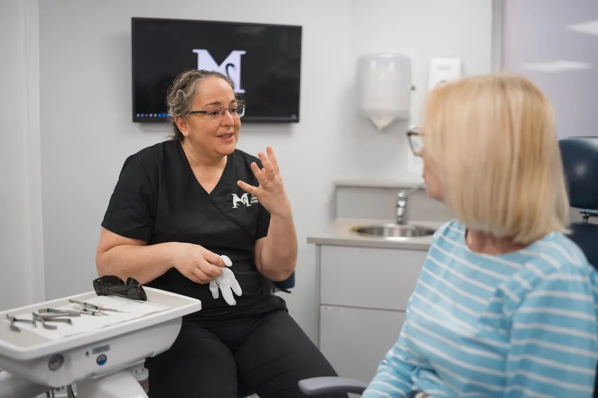 Dentist wearing black scrubs talks to a seated patient in a dental exam room.