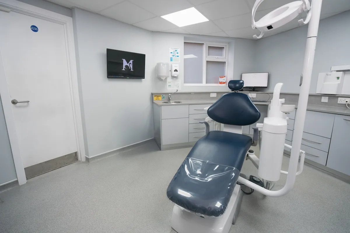 Modern dental clinic room with a black dental chair, monitors, and medical cabinets.
