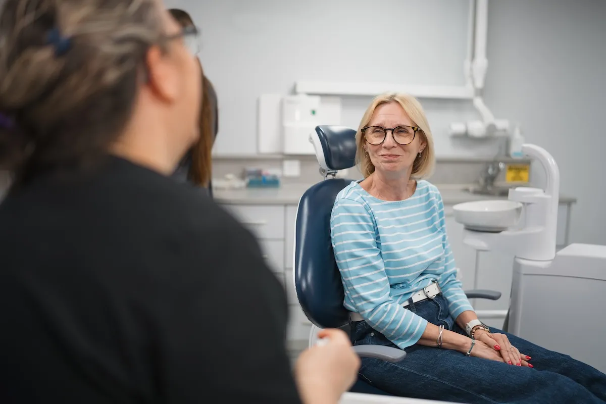 Smiling older woman with glasses in blue striped shirt sitting in a dental chair in clinic.