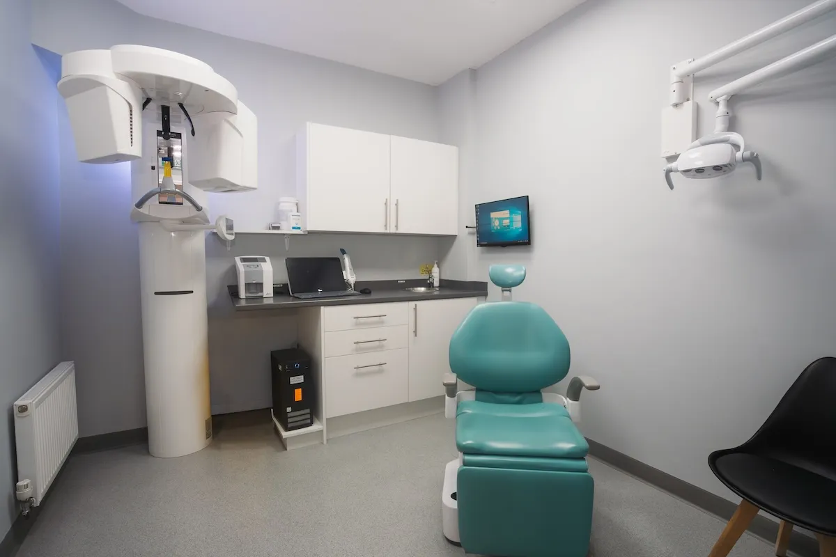 Modern dental examination room with teal chair, digital screen, white cabinets, and x-ray machine.