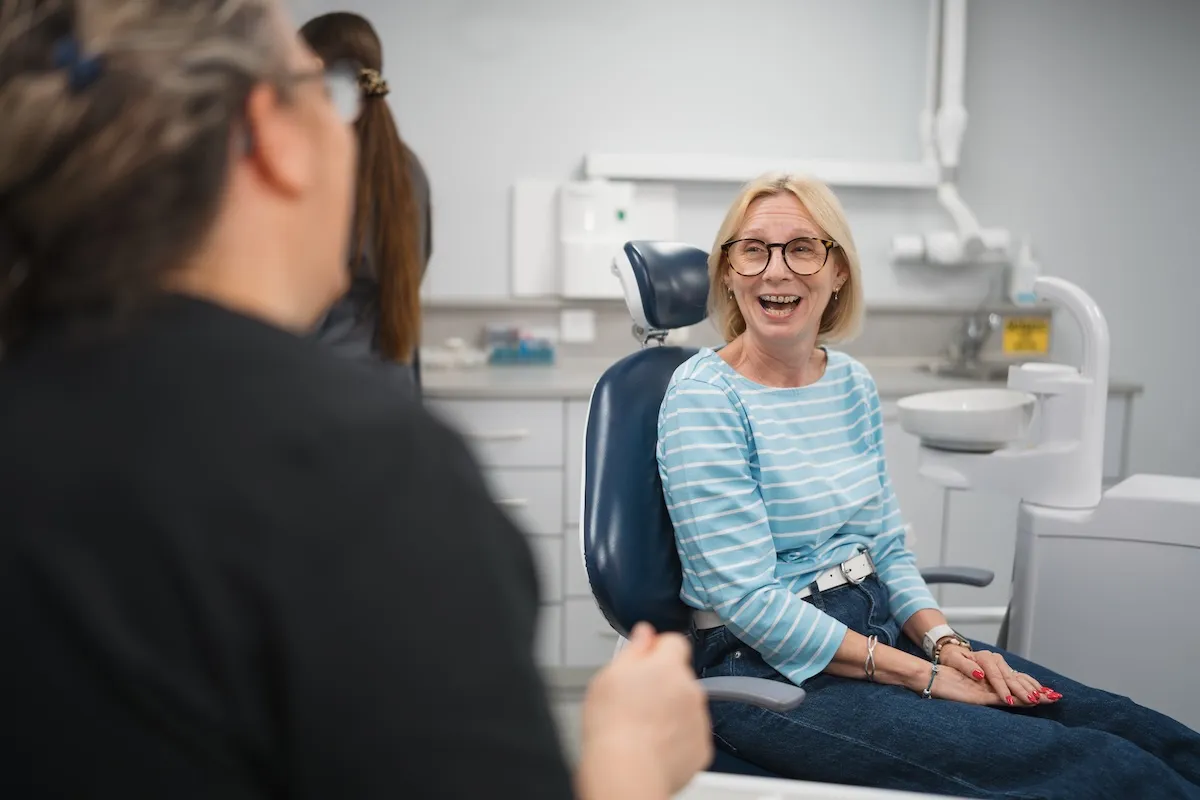 Smiling woman in glasses sitting in a dental chair talking to a person in black.