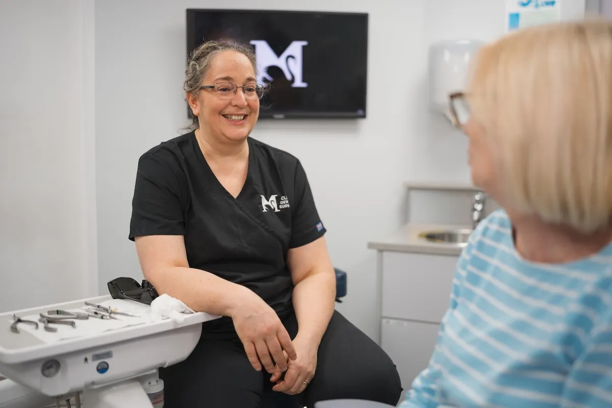 Smiling female dentist in black scrubs talks with a seated patient in a dental clinic.