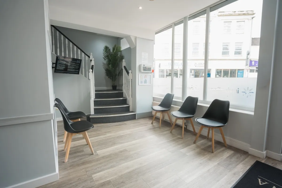 Waiting area with six black chairs near a window and stairs with a potted plant at the top.