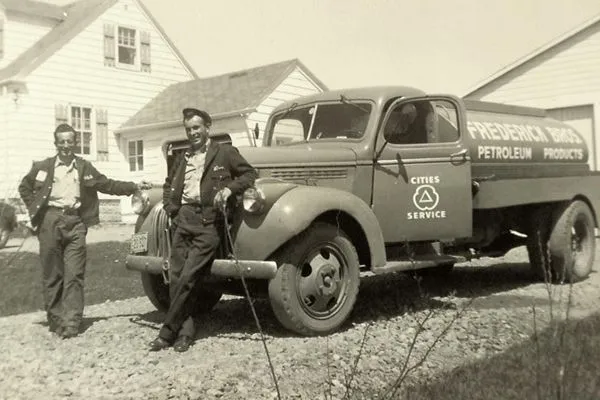 Black and white photo of two men standing beside a Fredericks Petroleum Products service truck parked in front of houses.