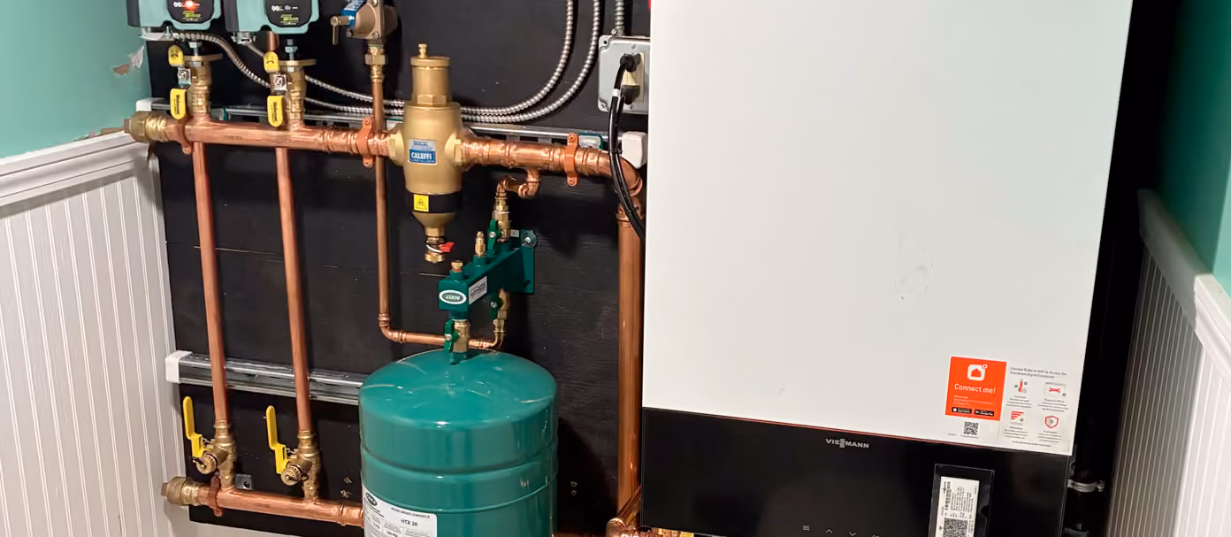 Propane heating installation with copper pipes, valves, and a wall-mounted boiler beside a green expansion tank.
